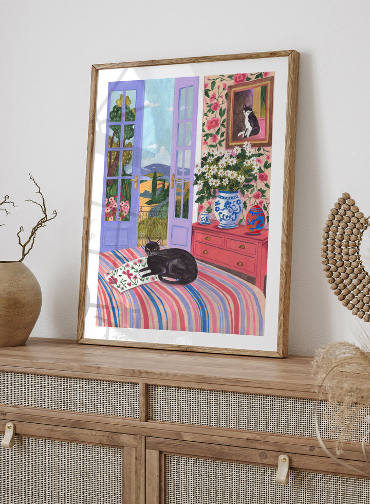 Cat lounging on a striped bed with a view of a garden through open doors, surrounded by flowers and decorative items. Black Cat on Bed French Doors Garden View by Caroline Bonne Müller is a colorful interior art print featuring cat illustration, floral room, and scenic garden view in a modern aesthetic style.