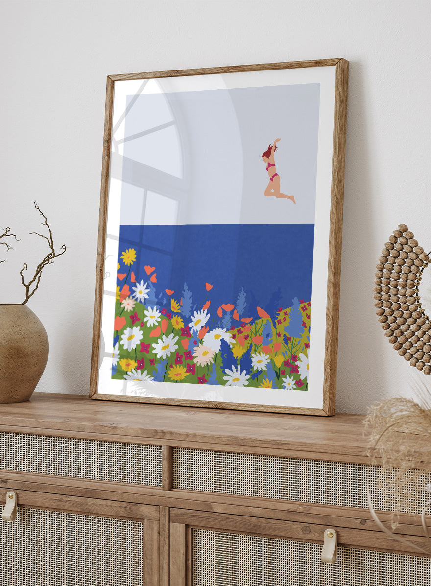 A framed art print featuring a minimalist illustration of a woman in a pink bikini jumping into a blue sea with a colorful wildflower meadow below. Wildflower Summer Jump art print by Ceyda Alasar features a joyful figure above colorful blooms, bringing playful summer energy to your walls.