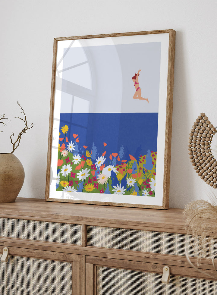 A framed art print featuring a minimalist illustration of a woman in a pink bikini jumping into a blue sea with a colorful wildflower meadow below. Wildflower Summer Jump art print by Ceyda Alasar features a joyful figure above colorful blooms, bringing playful summer energy to your walls.