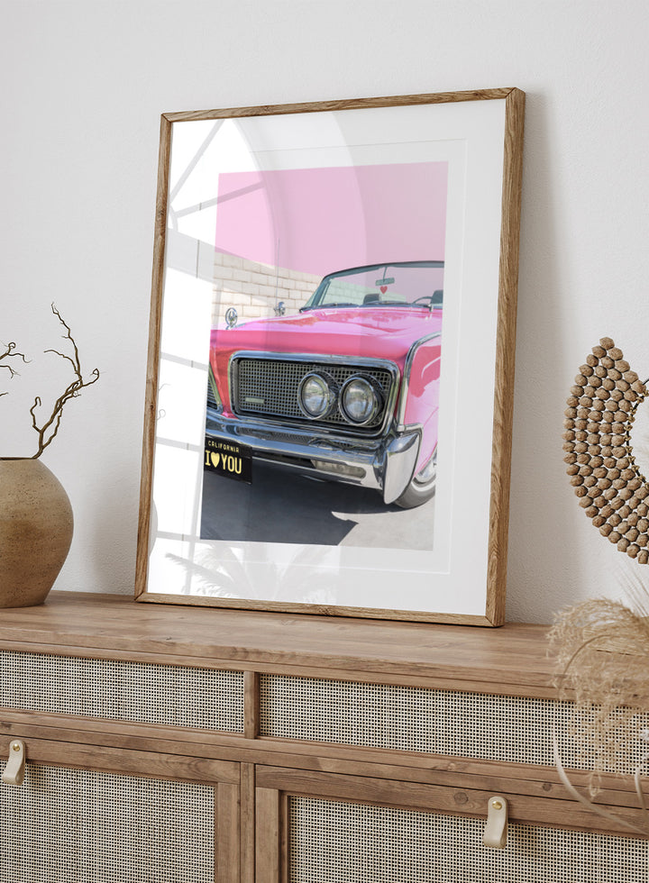 Pink vintage car with 'I ❤️ YOU' license plate against a pink sky. P.S. I Love You by Tom Windeknecht is officially licensed photography featuring a vibrant pop-art style photo of a car with romantic typography. Framed or unframed.