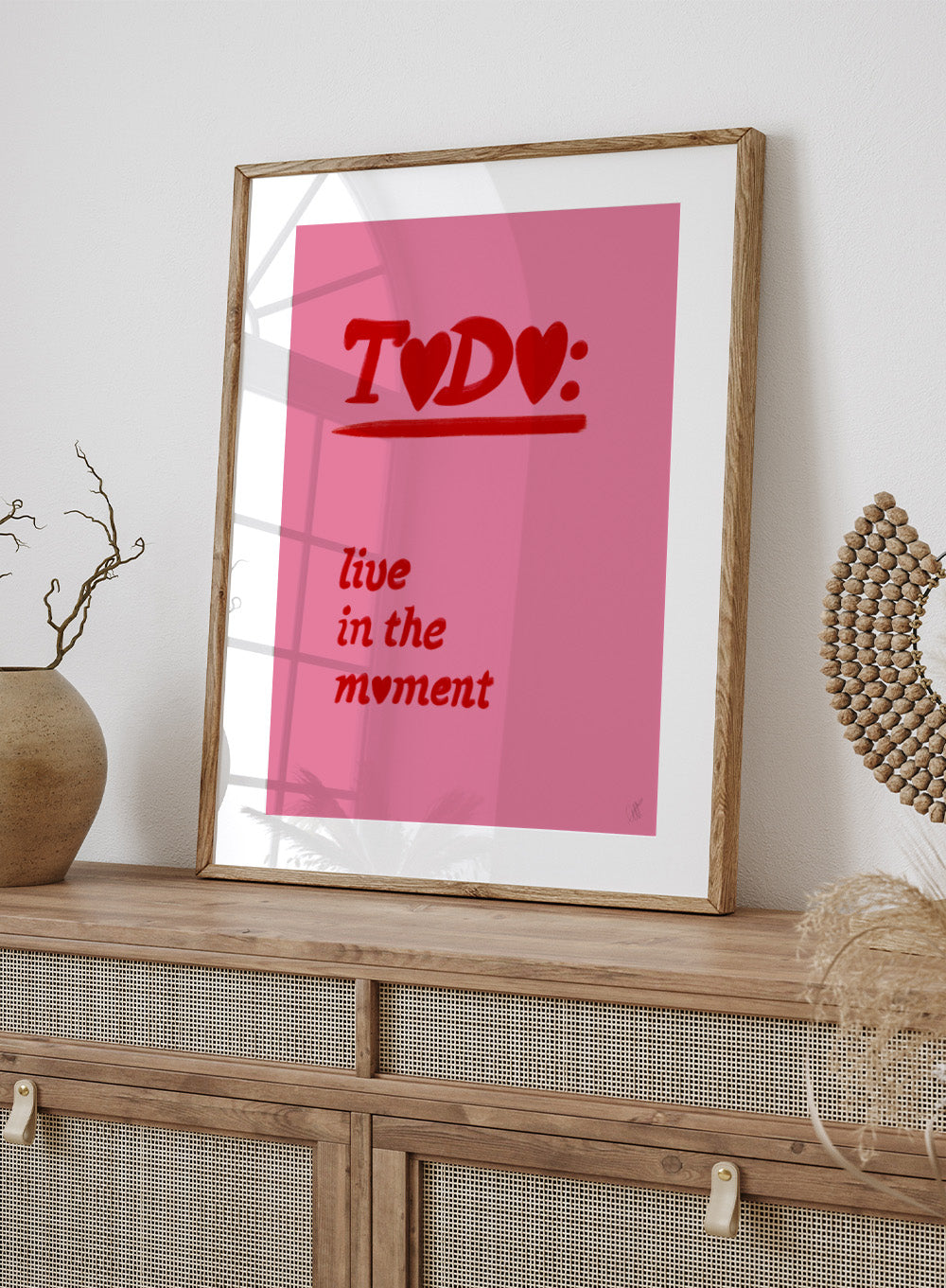 Pink poster with red text 'TODO: live in the moment' on a pink background. Typography wall art reading “live in the moment” with red hearts on pink background by Anne-Marie Volfova. Officially licensed motivational art print.
