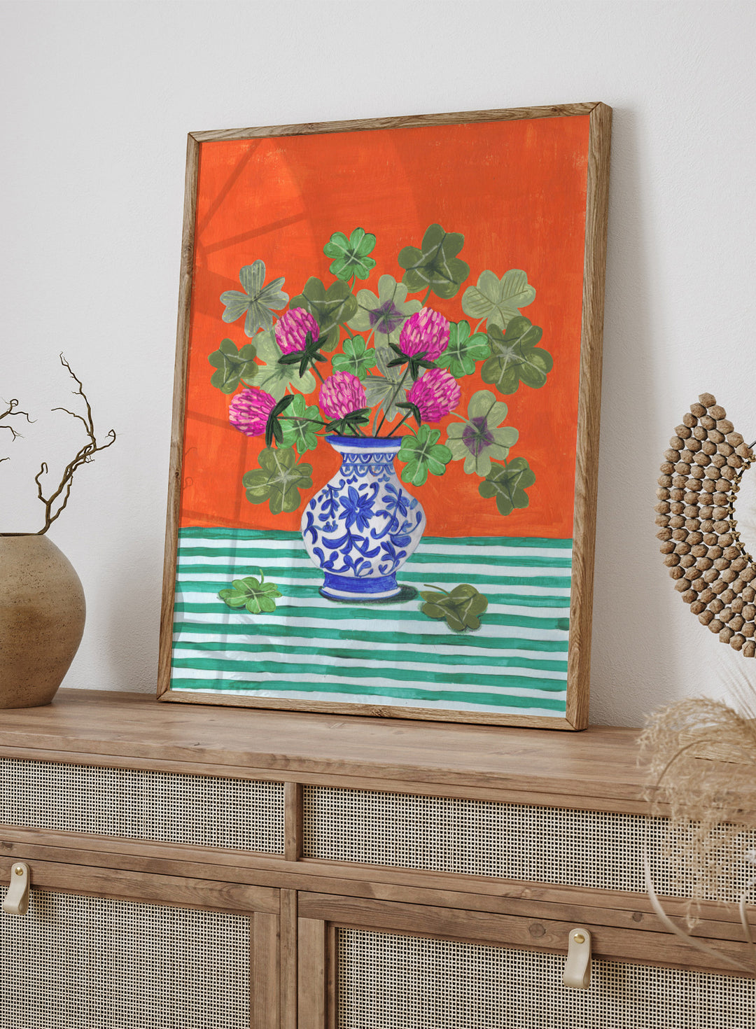 Vase with pink flowers and green leaves on a red and green striped background. Vibrant Clover Still Life by Caroline Bonne Müller is a fine art print and wall poster featuring clover leaves, pink flowers, and a blue vase on a striped table.