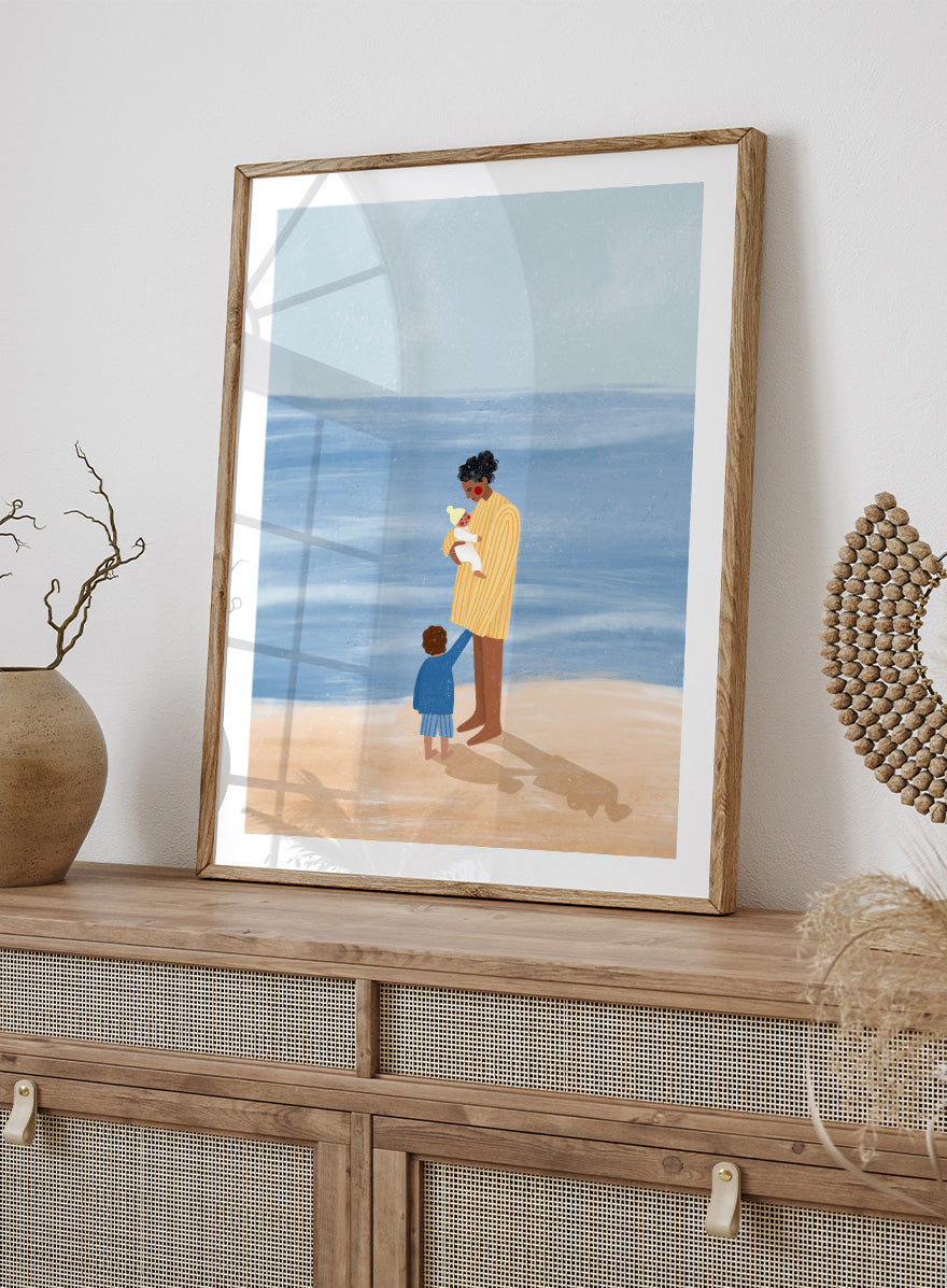 A framed art print featuring an illustration of a man in a yellow striped shirt holding a white dog on a beach next to a child in a blue coat, set against a light blue ocean and pale sky. Family Beach Day by Bea Müller art print featuring a mother with her baby and young child by the sea, created for warm family and coastal decor.
