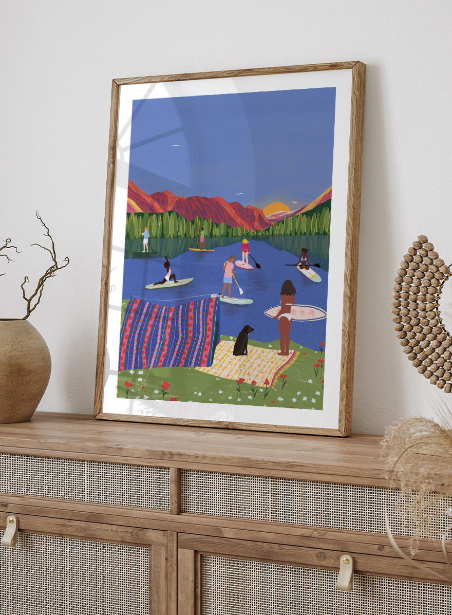 A framed art print featuring a colorful, stylized illustration of people paddleboarding and relaxing on a lake with mountains in the background. Paddleboard Lake Life by Ceyda Alasar outdoor art print featuring paddleboarders, mountain scenery, wildflowers, picnic blankets, and a lakeside dog.