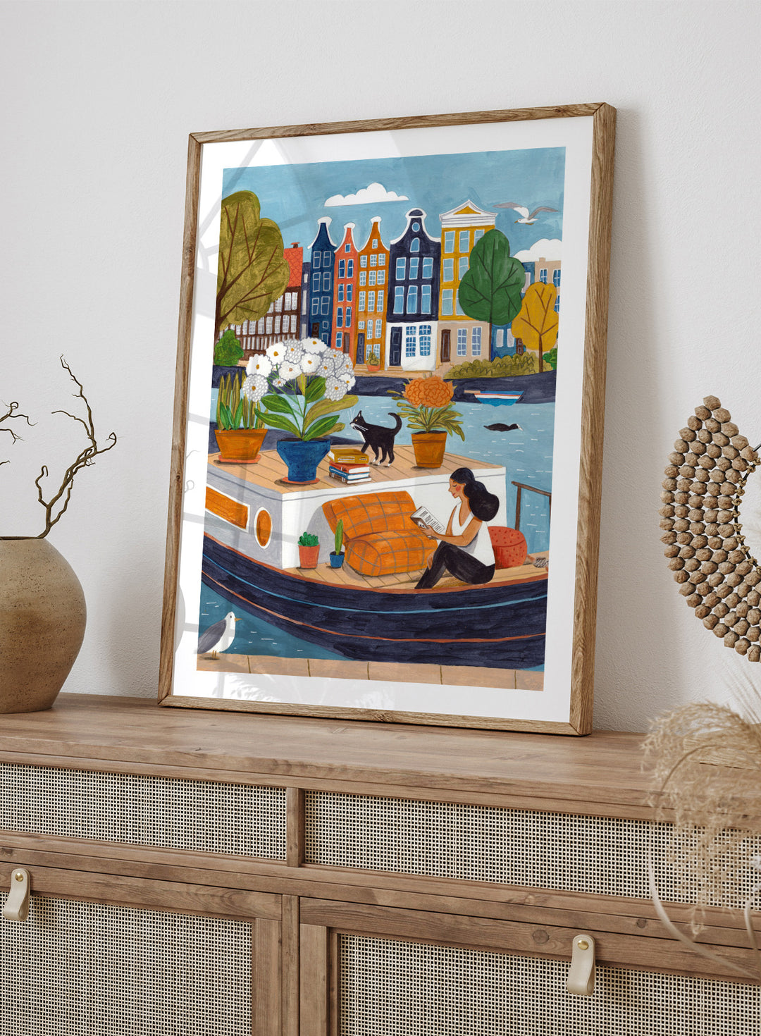 Illustration of a woman on a boat with flowers and a cat, surrounded by colorful houses. Cozy Canal Reading by Caroline Bonne Müller is a fine art print and wall poster featuring a woman reading on a canal boat with colorful European buildings in the background.