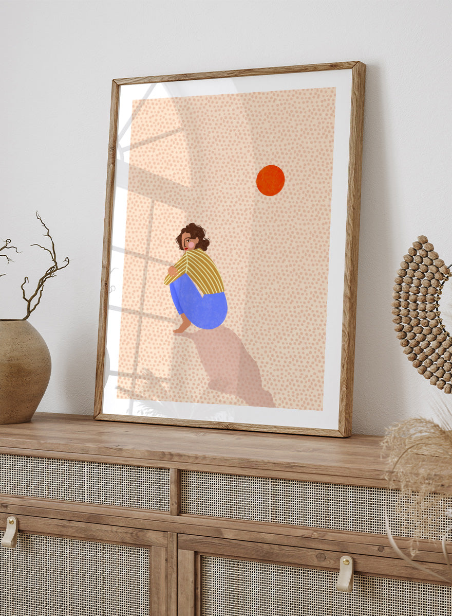 A minimalist art print featuring a stylized illustration of a person crouching in blue pants and a yellow striped top, looking up at a small orange sun against a light beige dotted background, framed with a white mat and black border. Pensive Woman Under Sun by Bea Müller art print featuring a seated woman beneath an orange sun in bold blue, mustard, and soft neutral tones.