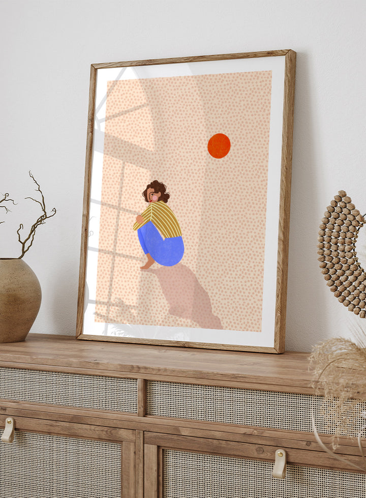 A minimalist art print featuring a stylized illustration of a person crouching in blue pants and a yellow striped top, looking up at a small orange sun against a light beige dotted background, framed with a white mat and black border. Pensive Woman Under Sun by Bea Müller art print featuring a seated woman beneath an orange sun in bold blue, mustard, and soft neutral tones.