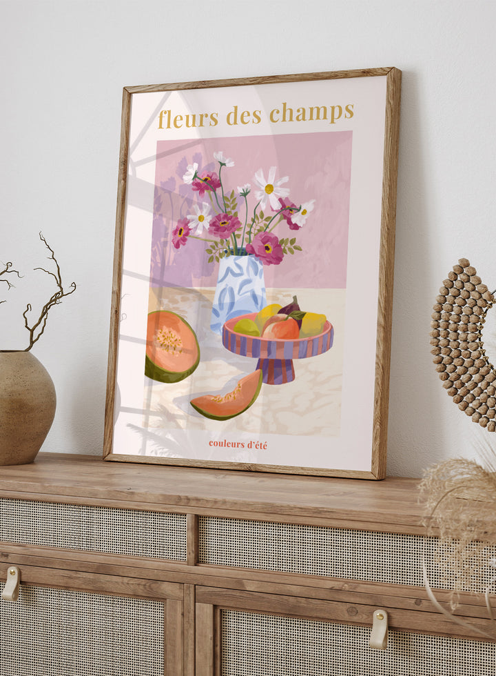 Illustration of a vase with flowers and fruits on a table against a pink background, with text 'fleurs des champs' and 'couleurs d'ete'. Fleur Des Champs Pink by Goed Blauw is a colorful still life art print and wall poster featuring flowers, fruit, and a patterned vase in a modern composition.