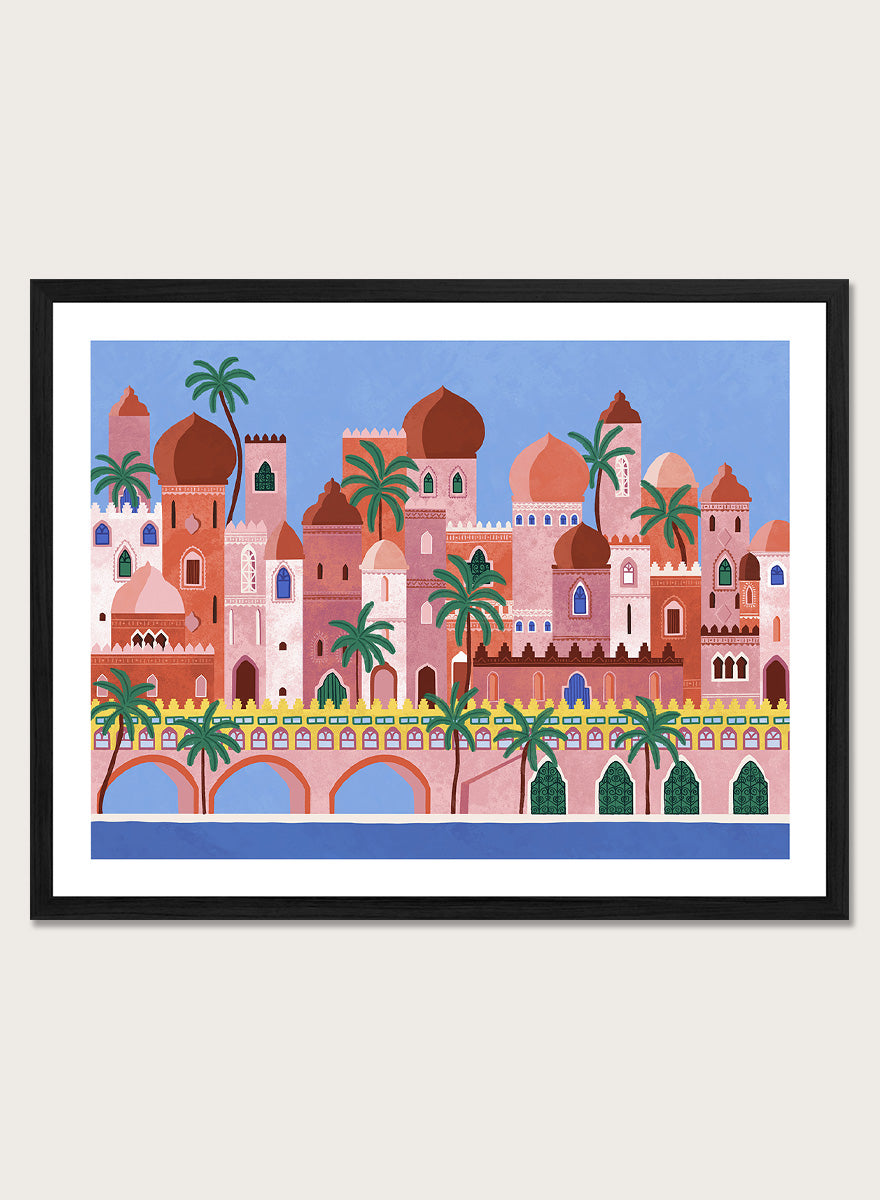 A framed art print featuring a stylized illustration of a coastal city with pink and peach buildings, green palm trees, and a blue sky. Arabian Fantasy City by Ceyda Alasar travel art print featuring ornate domes, arches, palm trees, patterned facades, and colorful desert architecture.