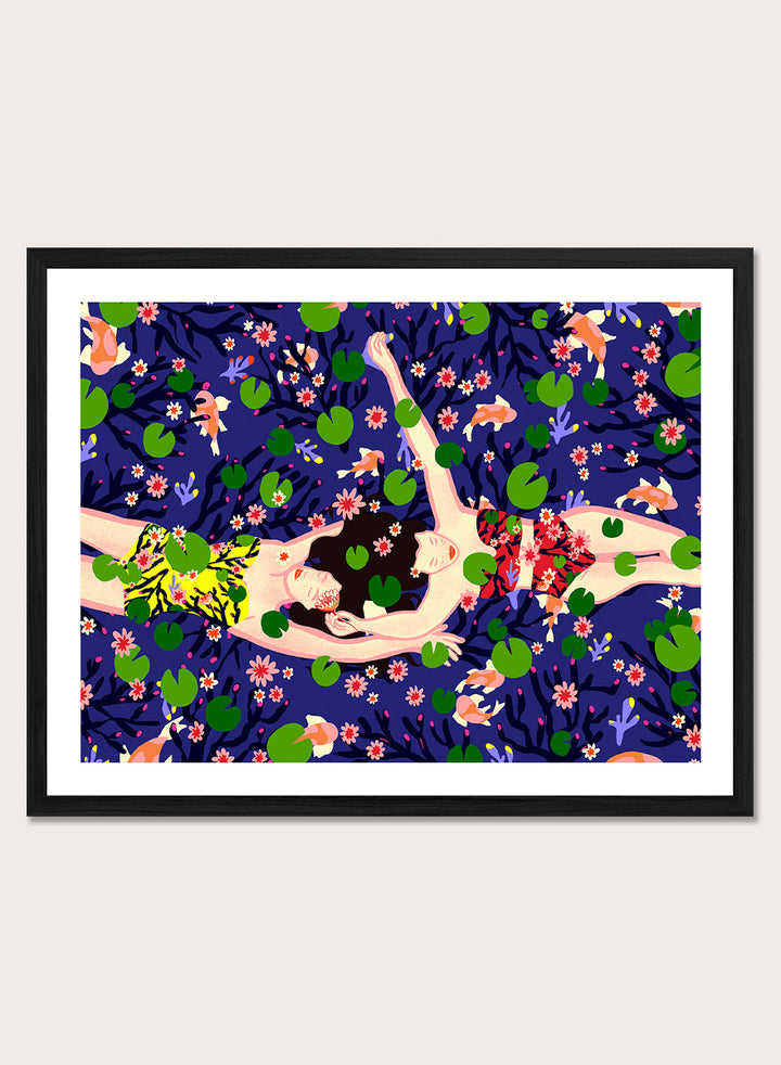 A framed art print featuring a stylized illustration of a woman lying on a dark blue background covered with green lily pads and pink flowers. Serene Night Float by Ceyda Alasar friendship art print featuring three women floating among lily pads, flowers, and fish in a deep blue pond.