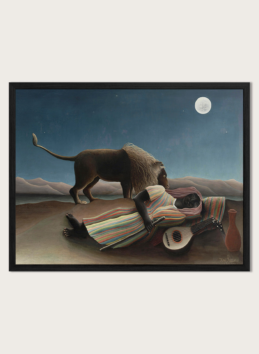 A framed surreal art print depicting a lion standing over a sleeping person wrapped in a striped blanket, with a guitar, vase, and full moon in a night sky. The Sleeping Gypsy by Henri Rousseau is a vintage art print featuring a sleeping traveler, lion, moonlit desert, striped robe, mandolin, and deep blue night sky.