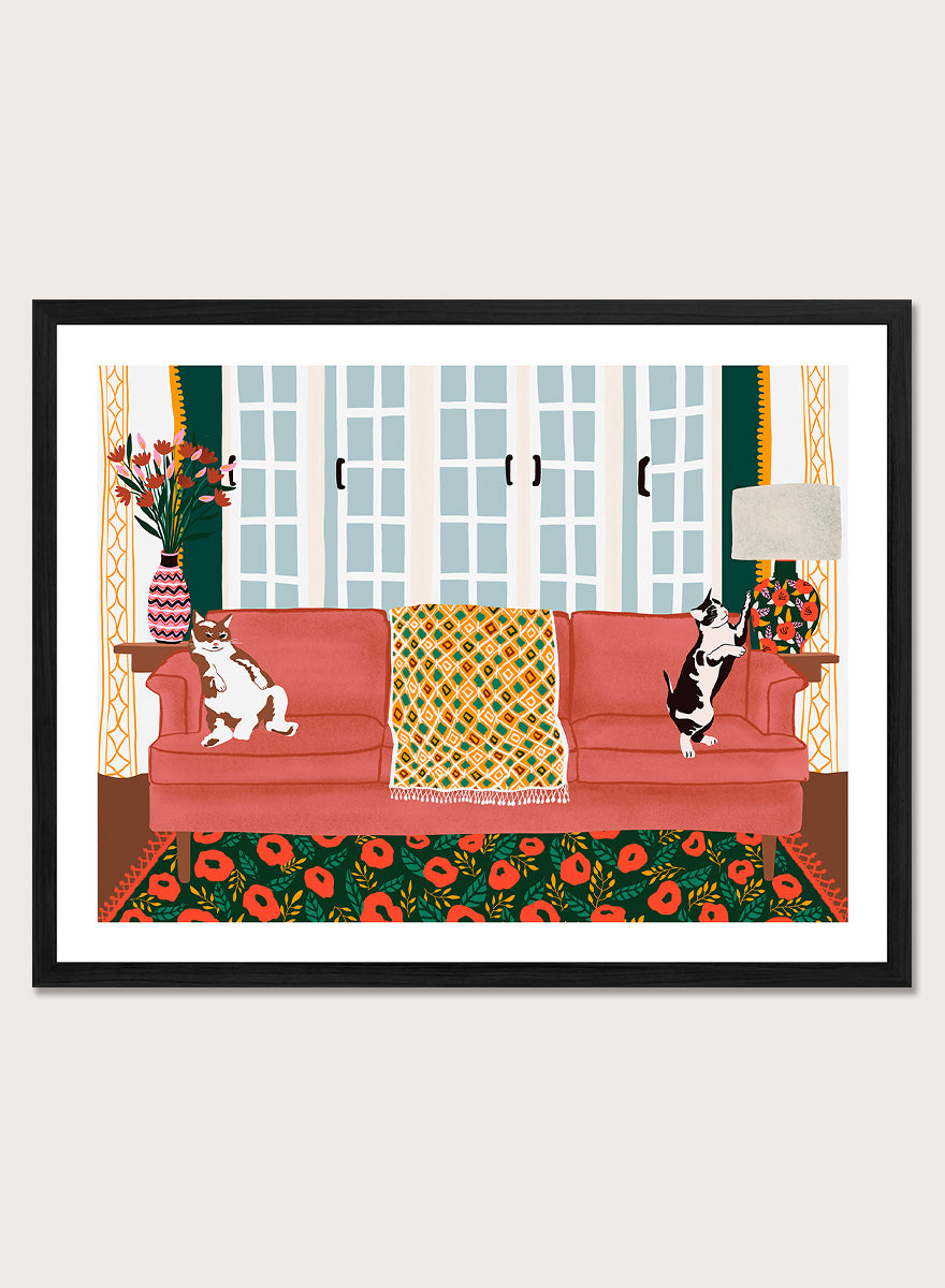 A framed art print depicting a cozy living room scene with a coral-pink sofa, two cats, a patterned throw blanket, and a floral rug. Playful Cats Living Room by Ceyda Alasar cat art print featuring two mischievous cats, a coral sofa, floral rug, patterned throw, and colorful decor.