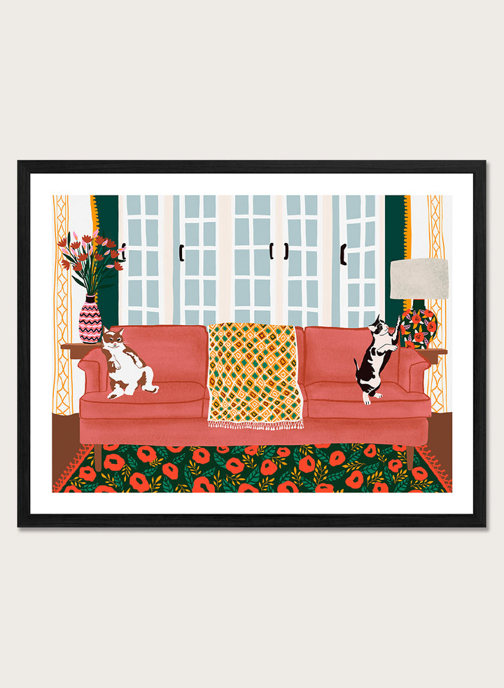 A framed art print depicting a cozy living room scene with a coral-pink sofa, two cats, a patterned throw blanket, and a floral rug. Playful Cats Living Room by Ceyda Alasar cat art print featuring two mischievous cats, a coral sofa, floral rug, patterned throw, and colorful decor.