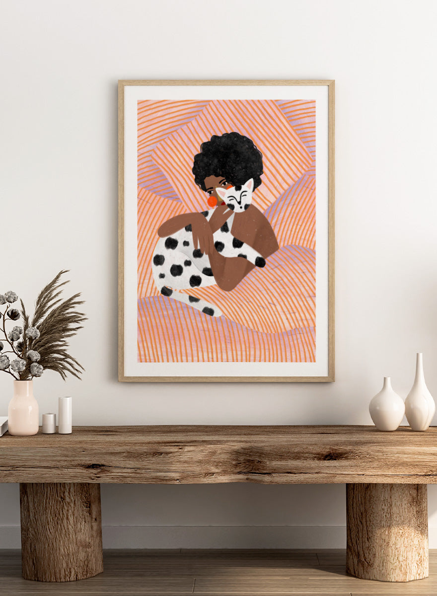 A framed art print featuring a stylized illustration of a person with an afro holding a black-and-white spotted cat, set against a peach and orange striped background. Cozy Cat Cuddles by Bea Müller animal art print featuring a woman embracing a spotted cat among soft peach and lilac stripes for cozy pet lover decor.