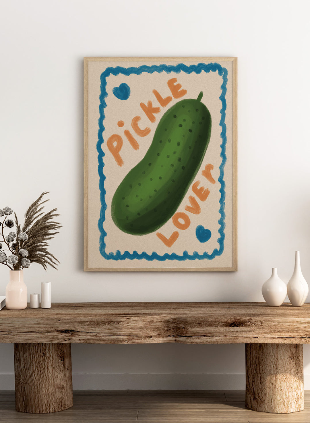 An art print featuring a hand-drawn green pickle centered on a cream background, surrounded by a blue scalloped border. The words &