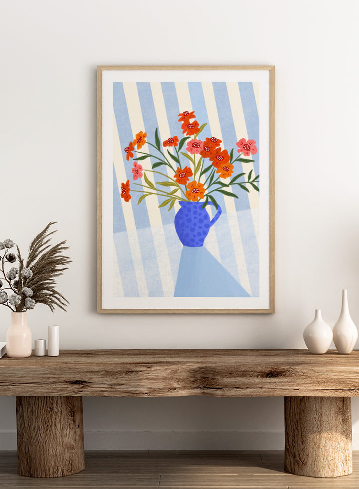 A framed art print featuring a stylized illustration of a blue patterned pitcher vase filled with orange, red, and pink flowers against a blue and white striped background. Cheerful Floral Pitcher by Bea Müller floral still life art print featuring orange and pink flowers in a bright blue jug with striped blue decor.