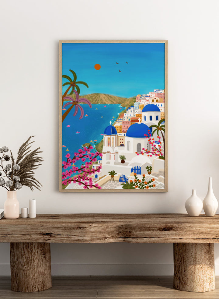 Shop Santorini Blue Domes by Nidhi Kachhadiya, a colorful Greek island wall art print inspired by Santorini, blue domes, whitewashed architecture, and Mediterranean views. A colorful illustrated coastal village scene with white buildings, bright blue domes, palm trees, pink flowering branches, a blue sea, and a clear sky with a sun and birds. 