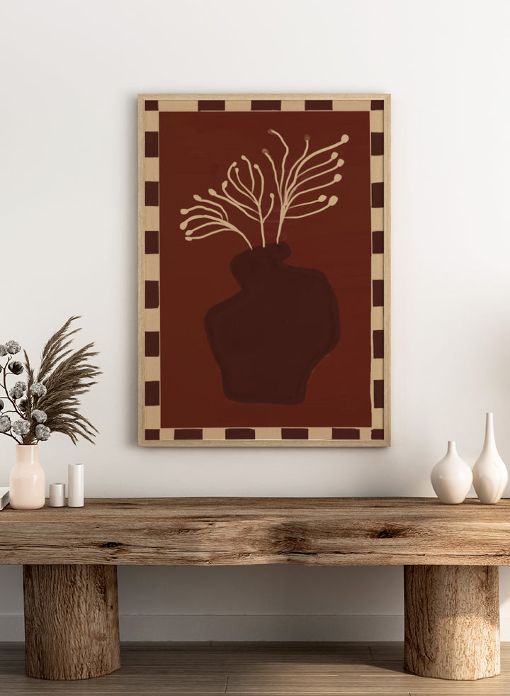 An art print featuring a dark brown abstract vase with cream branching stems on a deep reddish-brown background, framed by a beige and dark brown checkered border. Minimalist Vase Plant by The Art Concept still life art print featuring a dark vase, cream stems, checkerboard border, and warm rust-brown tones.