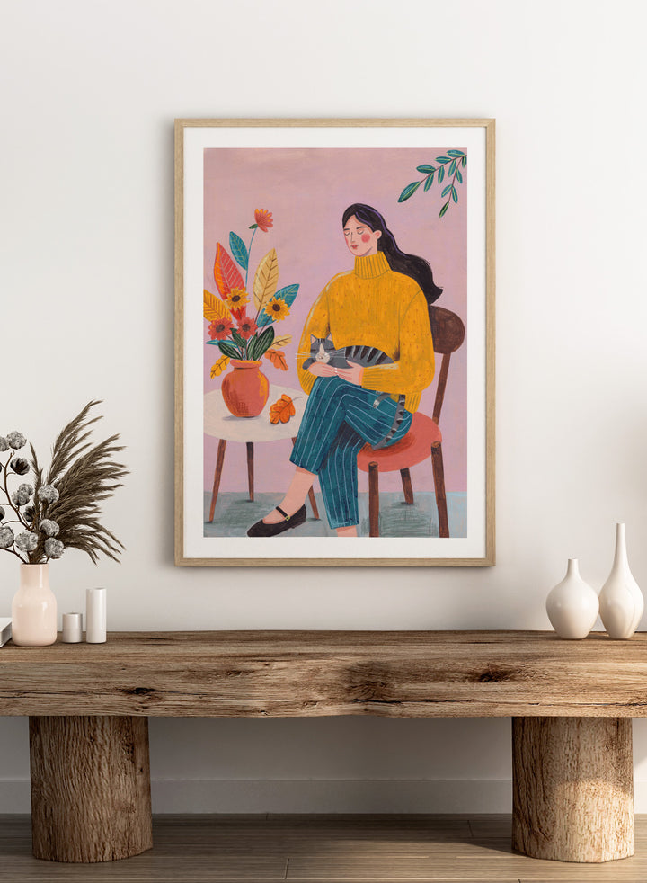 Illustration of a woman in a yellow sweater holding a cat, sitting by a table with flowers on a pink background. Peaceful Woman and Cat by Caroline Bonne Müller is a fine art print and wall poster featuring a woman holding a cat beside a floral arrangement.