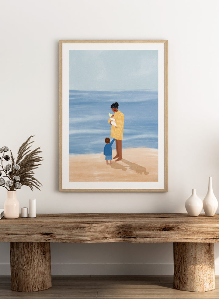 A framed art print featuring an illustration of a man in a yellow striped shirt holding a white dog on a beach next to a child in a blue coat, set against a light blue ocean and pale sky. Family Beach Day by Bea Müller art print featuring a mother with her baby and young child by the sea, created for warm family and coastal decor.