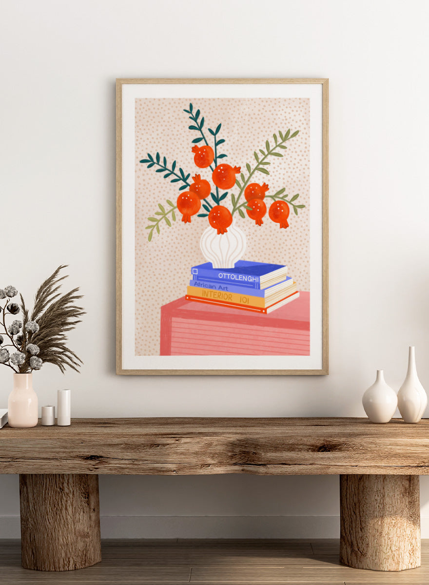 A framed art print featuring a stylized illustration of pomegranate branches in a white vase resting on a stack of books, set against a light beige dotted background. Cozy Tabletop Pomegranates by Bea Müller still life art print featuring red fruit branches, a sculptural vase, design books, and colorful interior styling.