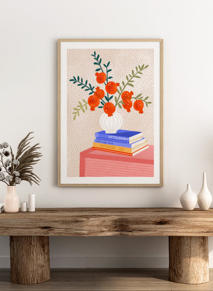 A framed art print featuring a stylized illustration of pomegranate branches in a white vase resting on a stack of books, set against a light beige dotted background. Cozy Tabletop Pomegranates by Bea Müller still life art print featuring red fruit branches, a sculptural vase, design books, and colorful interior styling.