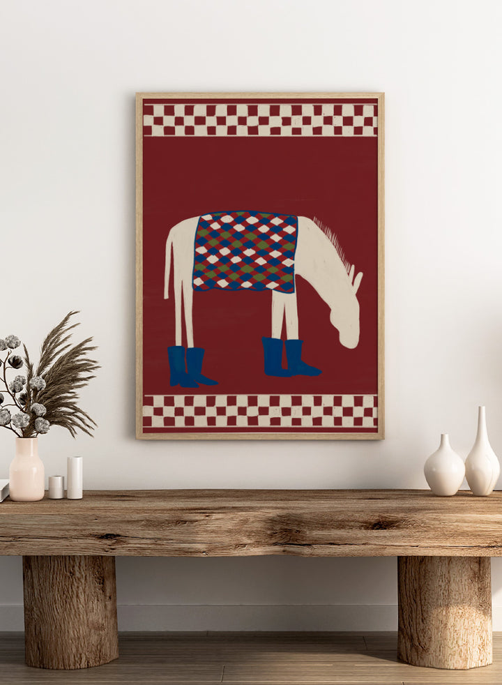 A stylized folk-art illustration of a white horse with a patterned blue, green, and red saddle blanket and blue boots, set against a dark red background with cream-and-red checkered borders at the top and bottom. Whimsical Donkey In Boots by The Art Concept animal art print featuring a white donkey in blue boots with a patterned blanket and rich red background.
