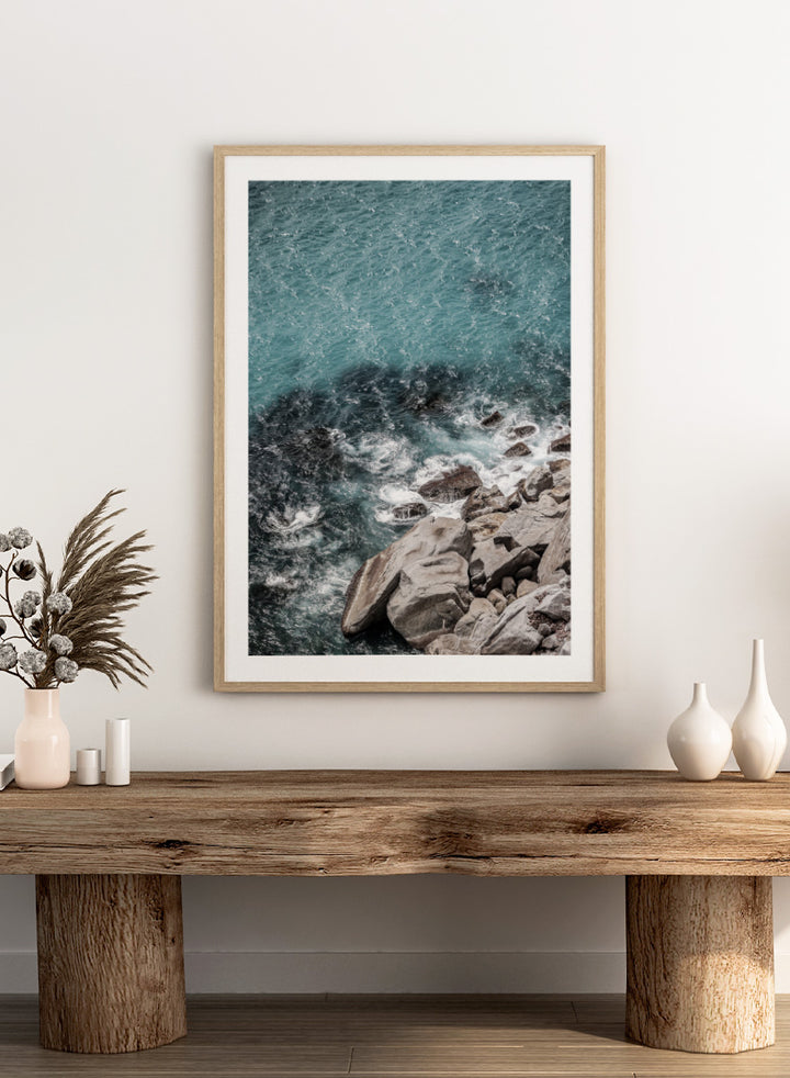 A framed photographic art print depicting a top-down view of turquoise ocean water washing over dark rocks and a rocky shoreline. Ocean’s Edge Symphony by Shot by Clint aerial ocean photography print featuring turquoise water, white surf, coastal texture, and soft seafoam tones.