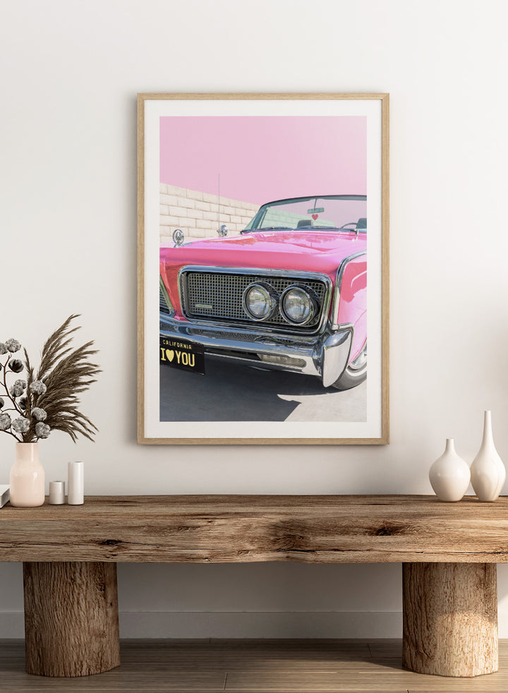 Pink vintage car with 'I ❤️ YOU' license plate against a pink sky. P.S. I Love You by Tom Windeknecht is officially licensed photography featuring a vibrant pop-art style photo of a car with romantic typography. Framed or unframed.
