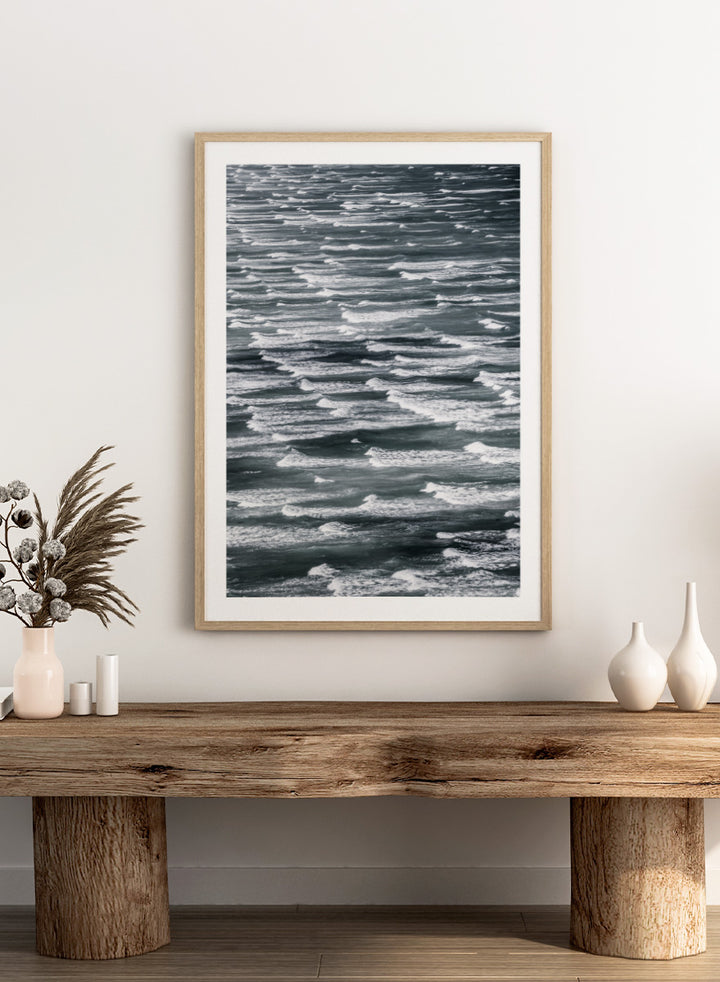 A framed black and white photographic print depicting a close-up view of ocean waves with white foam crests against dark water. Waves in Rhythm by Shot by Clint ocean photography print featuring layered blue-grey waves, white foam, and powerful natural movement across the sea.