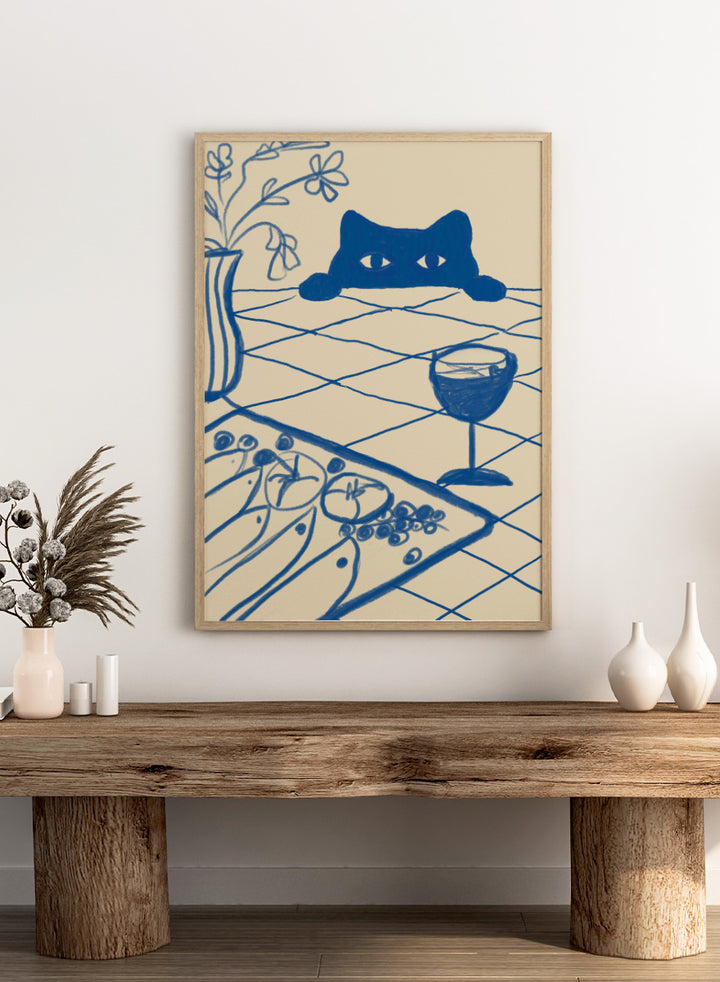 A stylized blue line-art illustration of a cat peeking over a table with a wine glass, flowers, and food, rendered in a cream background. Curious Blue Cat Dinner by The Art Concept is a playful cat art print featuring a peeking blue cat, table setting, flowers, and bold linework.