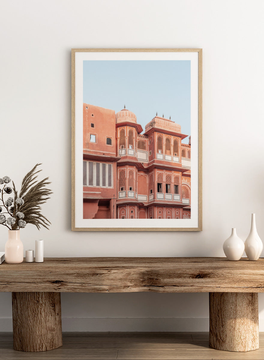 A framed art print featuring a photograph of the iconic pink sandstone Rajasthani palace, Udaipur, with white architectural details under a clear light blue sky. Terracotta Splendor by Shot by Clint architectural photography print featuring ornate palace windows, domed balconies, carved details, and warm coral tones.