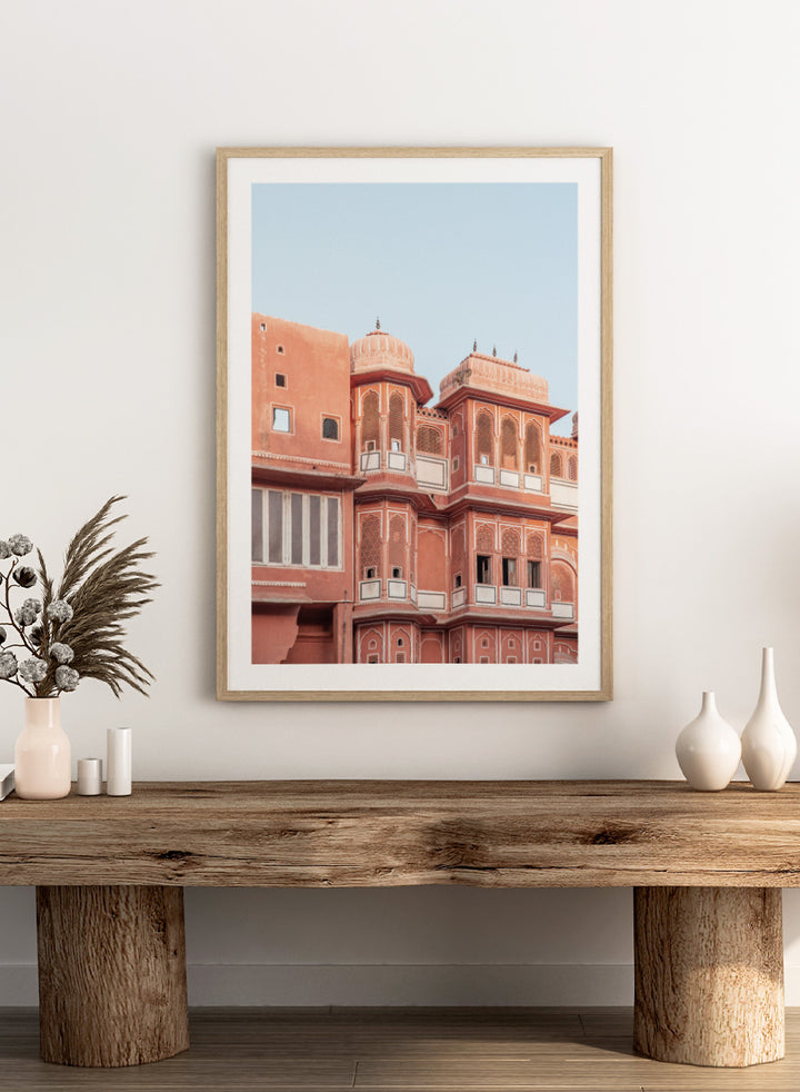 A framed art print featuring a photograph of the iconic pink sandstone Rajasthani palace, Udaipur, with white architectural details under a clear light blue sky. Terracotta Splendor by Shot by Clint architectural photography print featuring ornate palace windows, domed balconies, carved details, and warm coral tones.