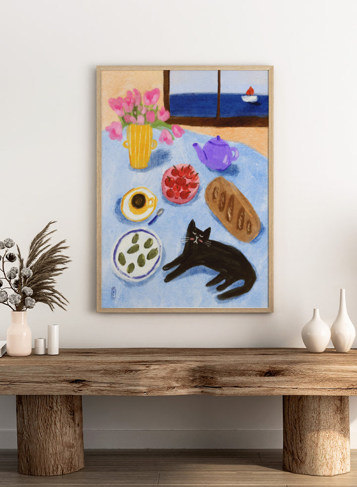 A colorful still life painting of a table scene with pink tulips in a yellow vase, a purple teapot, a bowl of strawberries, a cup of coffee, a loaf of bread, a black cat, and a window view of the sea with a small sailboat. Shop Relaxing Seaside Cat Breakfast by Dirkie Nell, a playful cat and food art print featuring breakfast, flowers, a sailboat, and a seaside view.