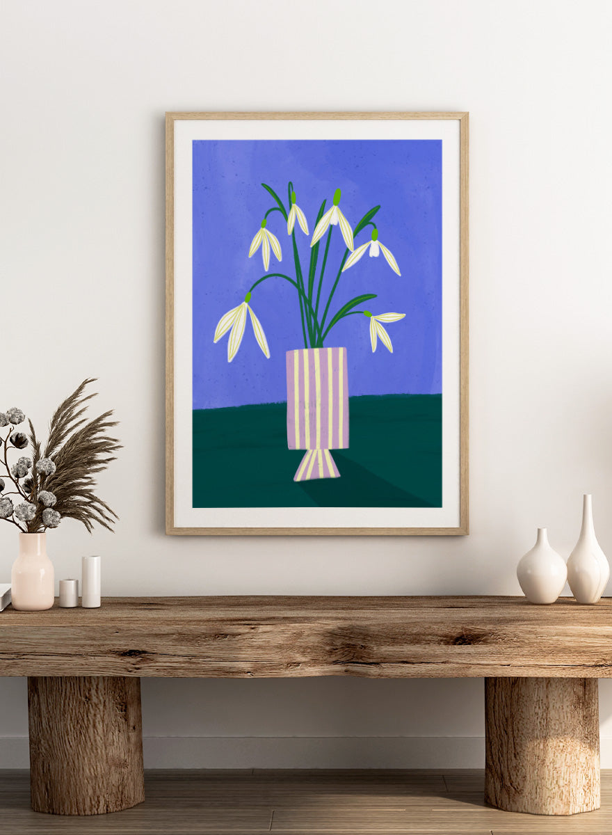 A framed art print featuring a stylized illustration of white snowdrops with green stems in a lavender and cream striped vase, set against a blue and dark green background. Snowdrops in Striped Vase by Bea Müller fine art print featuring white snowdrops, a lilac striped vase, and a deep blue background.