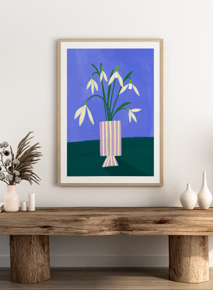 A framed art print featuring a stylized illustration of white snowdrops with green stems in a lavender and cream striped vase, set against a blue and dark green background. Snowdrops in Striped Vase by Bea Müller fine art print featuring white snowdrops, a lilac striped vase, and a deep blue background.