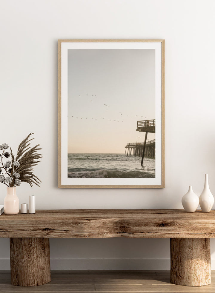 A framed photographic art print depicting a pier extending over a calm ocean under a pale sky, with birds flying across the scene. Seaside Serenity by Shot by Clint coastal photography print featuring ocean waves, a weathered pier, flying birds, and soft sunlit tones.