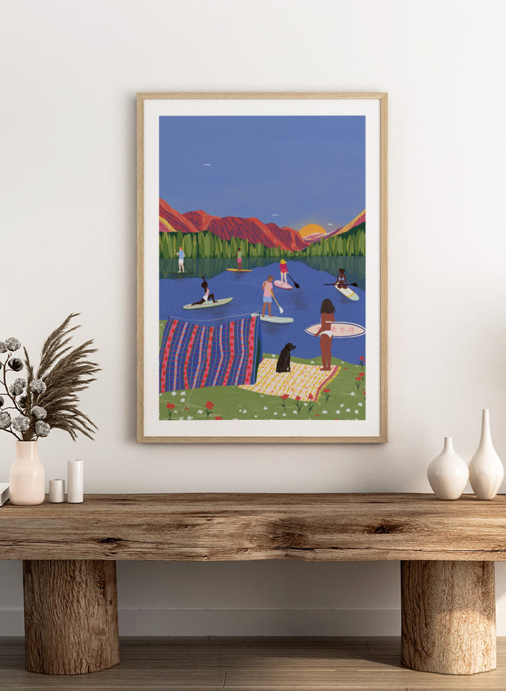 A framed art print featuring a colorful, stylized illustration of people paddleboarding and relaxing on a lake with mountains in the background. Paddleboard Lake Life by Ceyda Alasar outdoor art print featuring paddleboarders, mountain scenery, wildflowers, picnic blankets, and a lakeside dog.