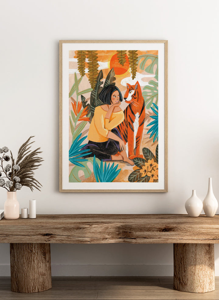 A framed art print featuring a stylized illustration of a woman in a yellow top and black skirt sitting beside an orange tiger, surrounded by tropical leaves and a warm orange sky. Woman with Tiger by Caroline Bonne Müller tropical art print featuring a woman beside a tiger among lush foliage, bold color, and hand-drawn texture.
