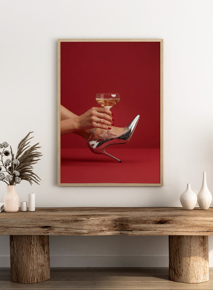 High-heeled shoe holding a glass of white wine against a red background, High Heels and Champagne by Andreas Magnusson is a fashion illustration art print featuring high heels, champagne glass imagery, bold red tones, and modern contemporary wall art style.