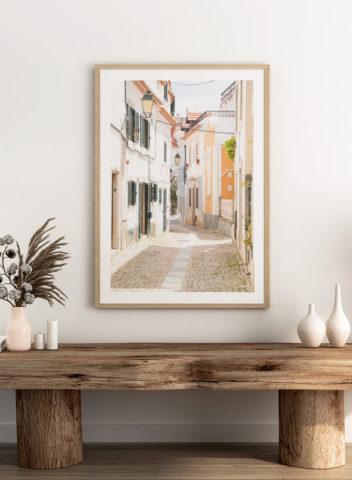 Cobbled street in a quaint European town with white buildings and a clear sky. Charming European Cobblestone Street by Henrike Schenk is a travel photography print featuring cobblestone streets, historic architecture, and European wall art.