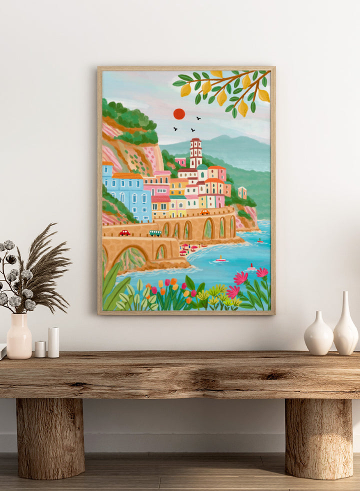 A colorful illustrated coastal town scene with pastel buildings, a stone bridge, blue water, mountains, flowers, and hanging yellow leaves. Shop Colorful Amalfi Coast by Nidhi Kachhadiya, a vibrant Italian coastal wall art print inspired by the Amalfi Coast, pastel villages, blue sea, and lemon trees.