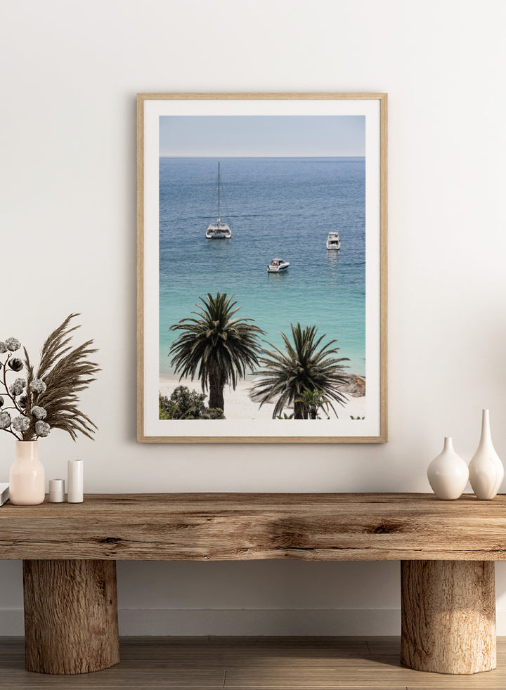 A framed coastal photograph showing a calm blue ocean with three boats, turquoise water near a white sandy shore, and two palm trees in the foreground. Tropical Serenity by Shot by Clint coastal photography print featuring turquoise water, sailboats, palm trees, and a bright tropical shoreline.