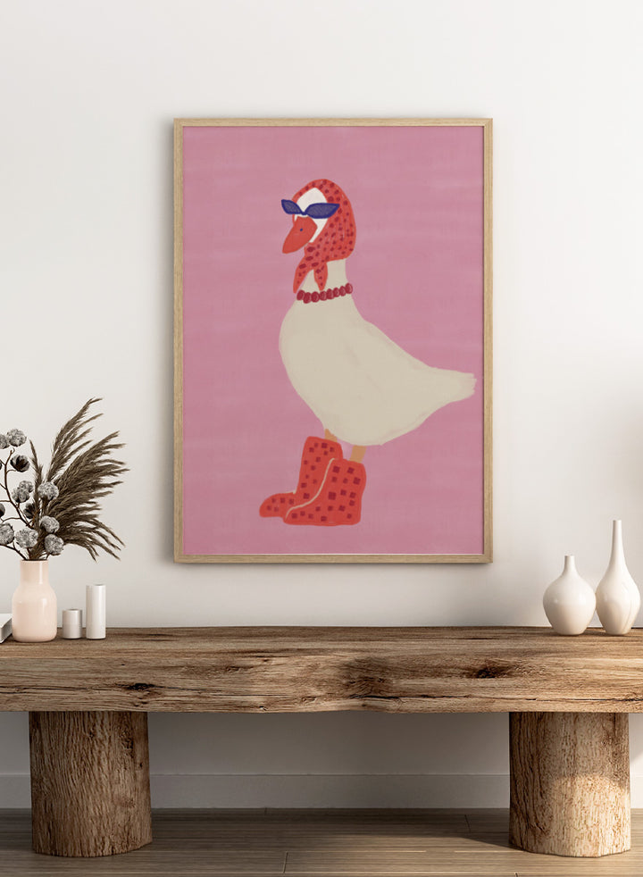 A stylized illustration of a white goose wearing a red patterned headscarf, blue sunglasses, a red beaded necklace, and red checkered boots, set against a solid pink background. Chic Goose Fashion by The Art Concept playful animal art print featuring a goose in sunglasses, a red headscarf, necklace, and bold red boots.