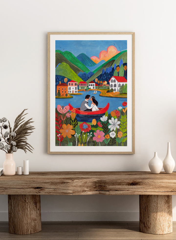 Couple in a red boat on a lake with mountains and houses in the background, surrounded by colorful flowers. Just Married Couple in a Boat by Caroline Bonne Müller is a romantic art print featuring couple illustration, lake scene, colorful flowers, and modern aesthetic wedding artwork.