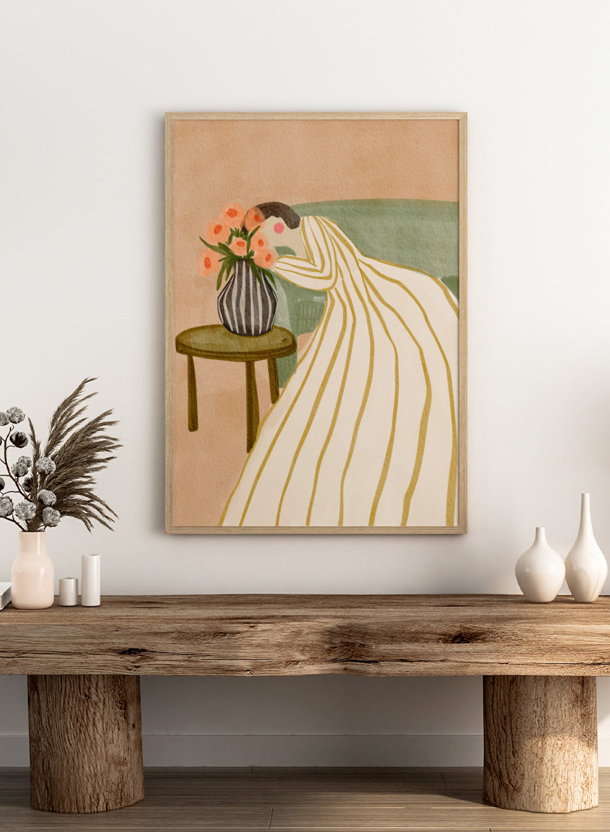 A stylized art print depicting a woman in a cream and gold striped gown leaning over a small table with a striped vase of pink flowers, set against a muted green sofa and beige background. Shop Woman Admiring Flowers by Nikita Jariwala, a modern floral wall art print featuring a woman, peach blooms, striped dress, and warm earthy tones.