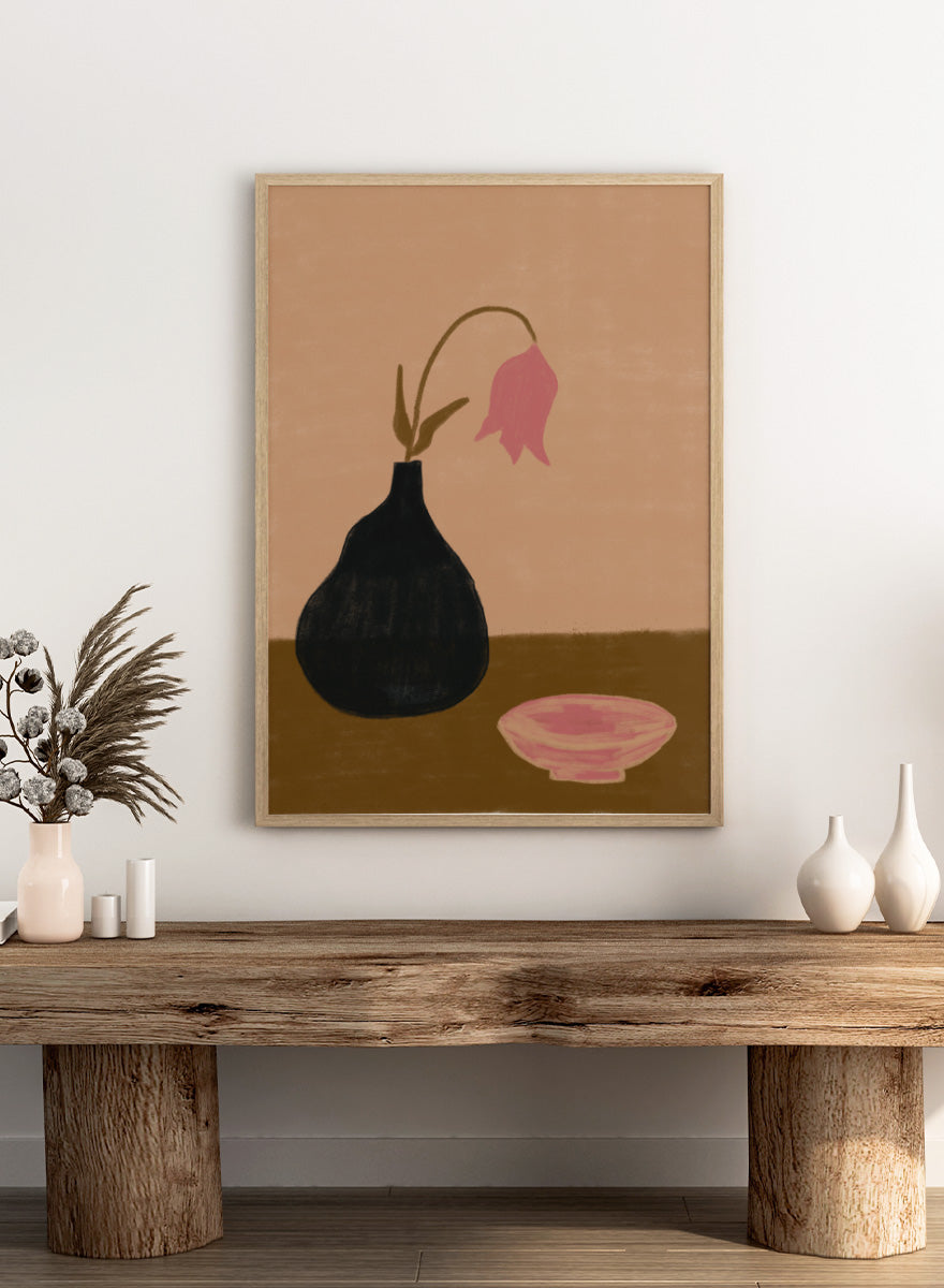 A minimalist still life painting featuring a black pear-shaped vase with a single drooping pink flower, alongside a pink bowl, set against a warm beige background and a brown tabletop. Minimal Pink Still Life by The Art Concept still life art print featuring a black vase, drooping pink flower, small bowl, and warm earthy tones.