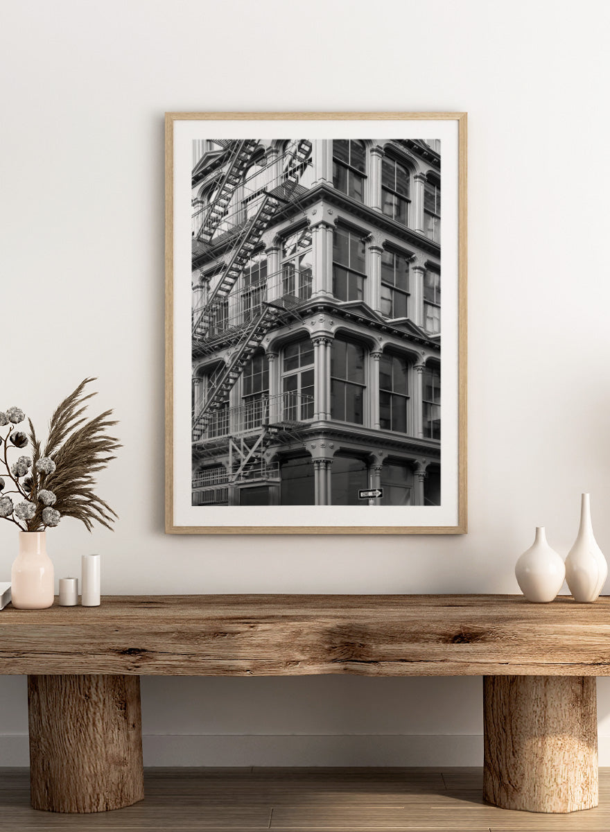 Black and white architectural photograph of a corner building facade with ornate windows and external fire escapes. Urban Geometry by Shot by Clint black-and-white architectural photography print featuring fire escapes, tall windows, ornate columns, and layered city detail.