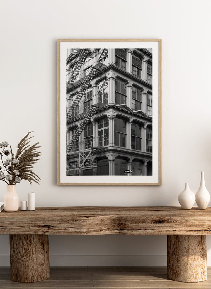 Black and white architectural photograph of a corner building facade with ornate windows and external fire escapes. Urban Geometry by Shot by Clint black-and-white architectural photography print featuring fire escapes, tall windows, ornate columns, and layered city detail.
