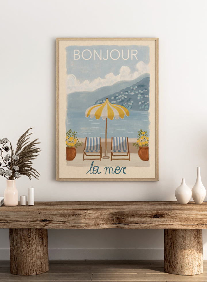 A vintage-style illustrated poster or print showing a beach scene with two striped lounge chairs under a yellow-and-white umbrella, potted plants, blue water, and mountains in the background. The words &